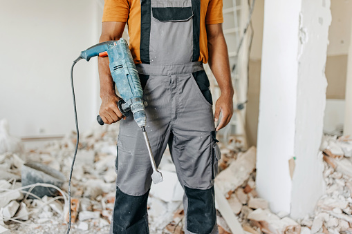 An African American male construction worker in protective uniform, holding a pneumatic sledgehammer in front of a room under renovation.