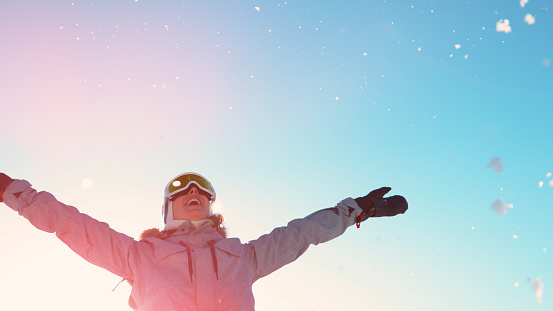 COPY SPACE, LENS FLARE: Happy female tourist plays with fresh snow in the sunny wintertime. Cheerful young woman snowboarding in the Slovenian mountains throws a handful of snow high in the air.