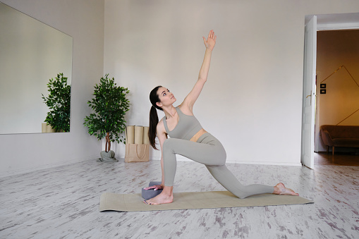 Side view of slim pretty positive young brunette woman doing Utthita parsvakonasana exercise, Extended Side Angle pose, on mat on floor surrounded by houseplants on white wall. Yoga and pilates