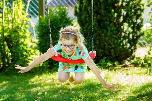 Happy little positive preschool girl having fun on swing in domestic garden. Smiling child with glasses swinging on sunny day. Active leisure and activity outdoors. stock photo