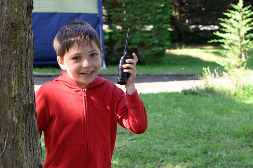 Portrait of a kid looking at the camera. The boy is holding a walkie talkie in his hand and is in a camping site