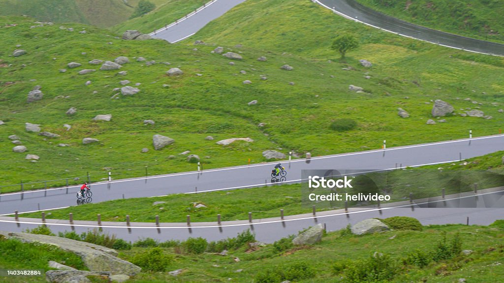 Two active tourists ride their bikes down wet switchback road in Swiss mountains Two active tourists ride their bikes down a wet switchback road high in the picturesque mountains of Switzerland. Unrecognizable bicycle riders cruise down the Furka pass road on a rainy summer day. Furka Pass Stock Photo