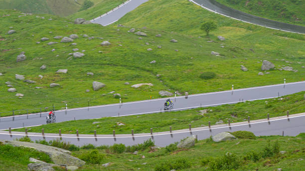 Two active tourists ride their bikes down wet switchback road in Swiss mountains Two active tourists ride their bikes down a wet switchback road high in the picturesque mountains of Switzerland. Unrecognizable bicycle riders cruise down the Furka pass road on a rainy summer day. furka pass photos stock pictures, royalty-free photos & images