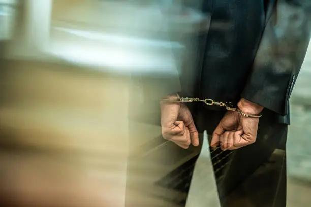 Hand close-up of unrecognizable businessman in handcuffs getting arrested in the office. Shot through glass. Online harassment, hacking, digital theft.