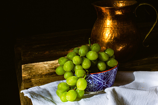 a blue bowl holding a mound of green grapes on a linen napkin in front of a copper jug on a rustic table