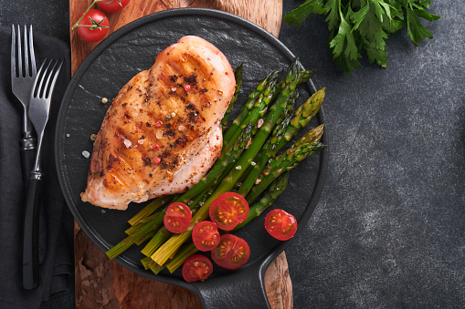 Chicken breast grilled with vegetables. Grilled chicken steak, asparagus and cherry tomatoes in black plate on gray concrete table background. Healthy diet lunch. Barbecue steak fried. Top view.