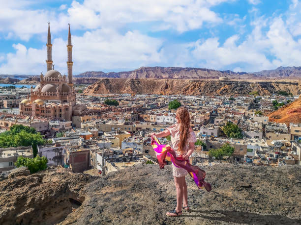 Blonde-haired young woman dressed in light summer clothes and sandals looks at the Al Sahaba Mosque in Sharm El Sheikh from a mountain top, Egypt stock photo