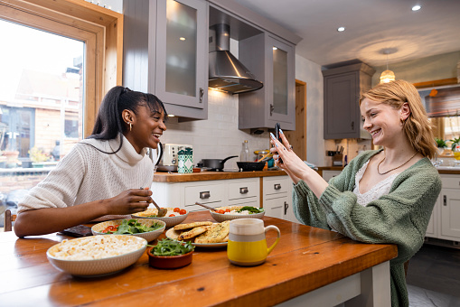 Side-view shot of two friends maintaining their sustainable living by enjoying a vegan meal at their home in the North East of England. They are eating and talking, one young woman is taking a picture of her friend.