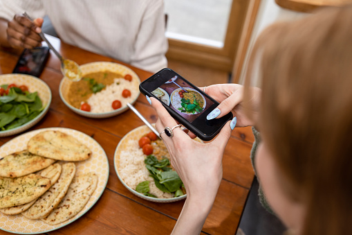 A close-up high angle view shot of an unrecognisable woman having a meal with her friend, they are maintaining their sustainable living by enjoying a vegan meal at their home in the North East of England. One young woman is taking a picture of the food with her smartphone.