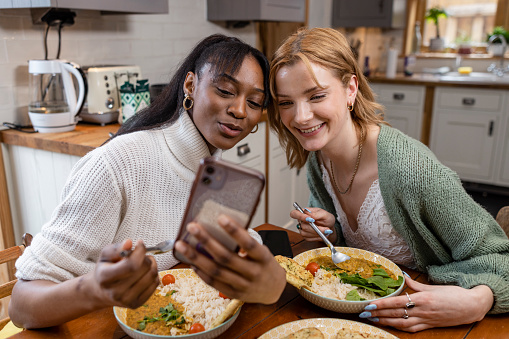 Two friends maintaining their sustainable living by enjoying a vegan meal at their home in the North East of England. They are taking a selfie together on a smartphone.