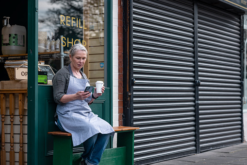 Woman working at a cafe/store that promotes sustainable living in the North East of England. The store has refill stations to reduce plastic and food waste. She is wearing an apron and sitting down outside the front of the store to enjoy her break with a coffee and a phone call on her smart phone.