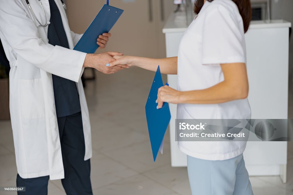 Close up of two doctors in uniform shaking hands standing in clinic hall near reception Close up of two doctors shaking hands standing in clinic hall near reception. High quality photo Medical Exam Stock Photo