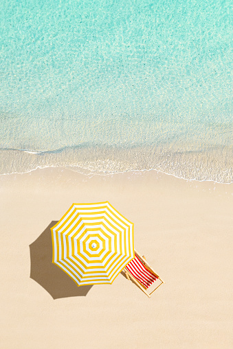 Top view of sunbed, lounge, yellow umbrella, SUP board for surfing on the shore on tropical Seychelles sand beach. Blue, turquoise transparent water surface of ocean, sea, lagoon. Aerial, drone view. vertical