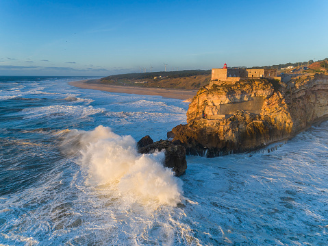 Aerial view of lighthouse on a cliff with a fortress on the coast of the Atlantic ocean with big waves at sunset in Nazare, Portugal