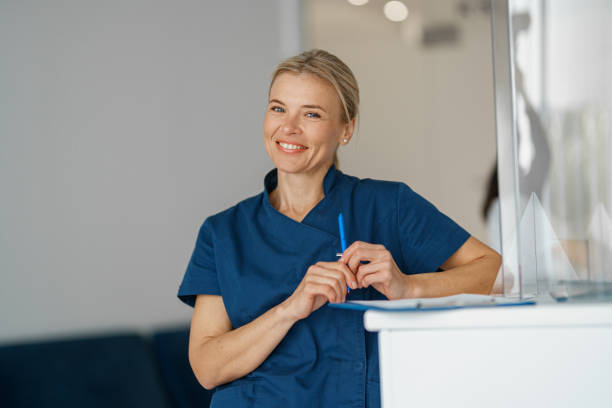 Smiling doctor with clipboard standing near reception in clinic hall and looking camera stock photo