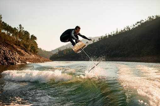 young active man in black wetsuit masterfully jump with foil wakeboard over splashing wave. Water sports activity.
