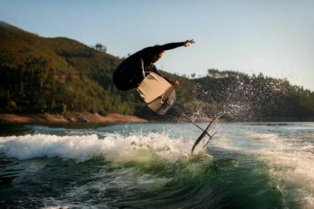 energy athletic man in black wetsuit jump with foil wakeboard over splashing wave. Water sports activity.