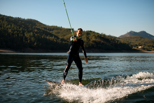 active male rider in black wetsuit holds rope with handle and riding on wakesurf board on splashing water.