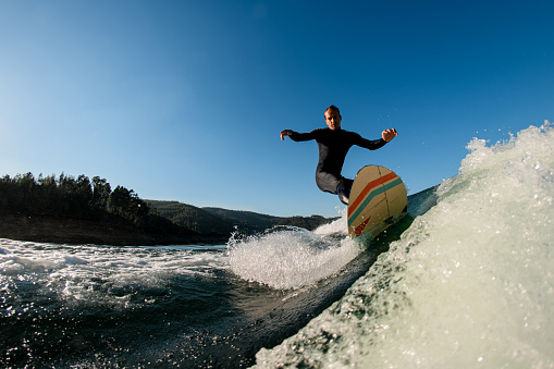 man wakesurfer in a black wetsuit riding down the splashing wave on blue sky background