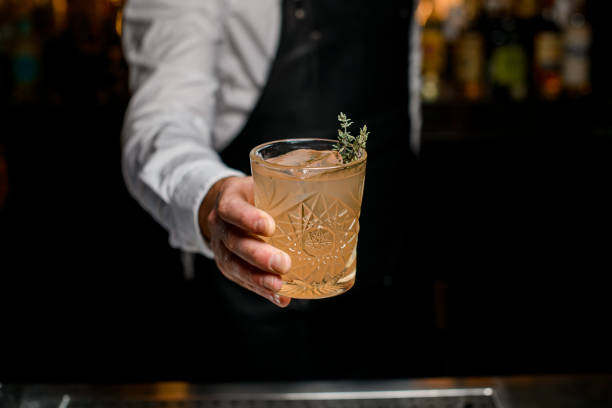 great view of glass with cocktail garnished with rosemary in the hand of bartender on blurred background - transparent holding glass focus on foreground imagens e fotografias de stock