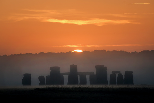 The solstice sun breaks at dawn  over the prehistoric Stonehenge monument in Wiltshire, England. Distant silhouette of the ancient monument the morning mist
