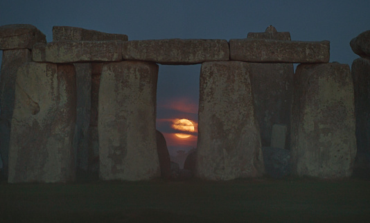 The full moon descends as its sets at dawn  behind  the prehistoric Stonehenge monument in Wiltshire, England. The setting full moon glows through the ancient stones