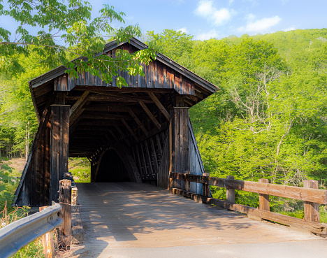 Weathered wooden country covered bridge, seen from entrance on a summer day. Trees and grass surrounding and blue sky above. Bucolic, serene country mood. Selective focus on bridge structure.