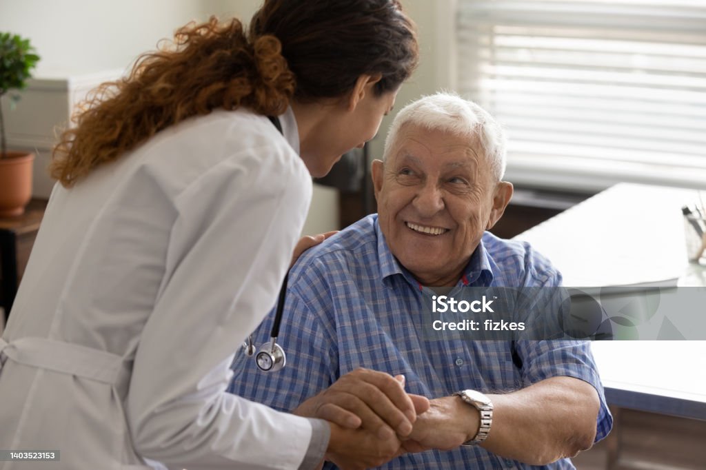 Friendly doctor and optimistic elder 80s man meeting in office Friendly young female doctor and optimistic elder 80s man meeting in office for consultation. GP therapist holding hands of old patient, giving sympathy, support, advice about treatment Senior Adult Stock Photo