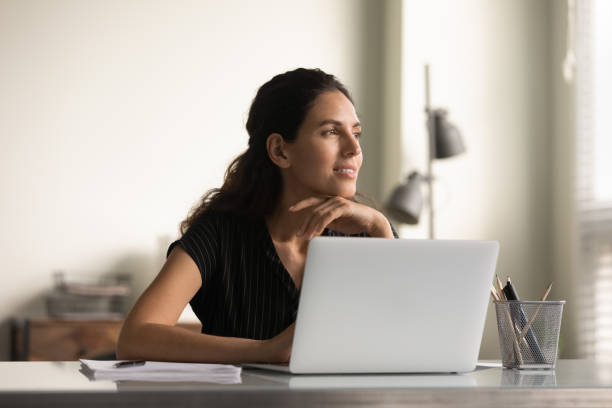 улыбающаяся вдумчивая сотрудница, работающая за ноутбуком из дома - decisions women thinking latin american and hispanic ethnicity стоковые фото и изображения