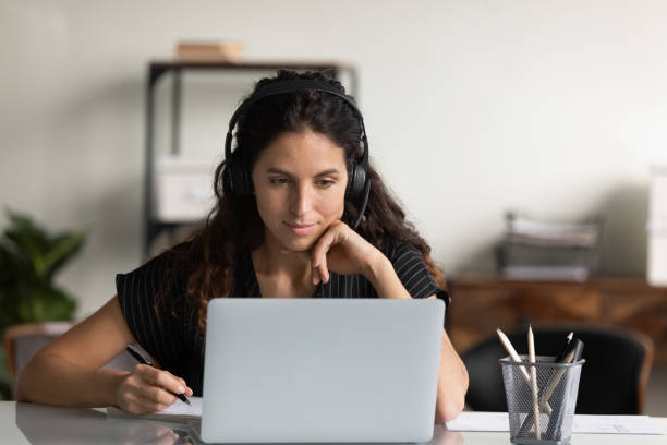 Focused female adult student in headphones using laptop Focused female adult student in headphones using laptop, attending online lesson, virtual class, listening webinar, watching training, writing notes, making video call. Remote learning on internet business teaching student writing stock pictures, royalty-free photos & images