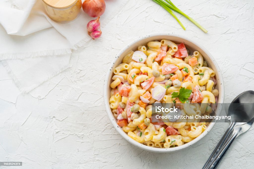Homemade macaroni salad with elbow pasta Homemade macaroni salad with elbow pasta, onion, carrot, tomato, green peas and mayonnaise dressing in a white bowl on a white wooden table.Top view Salad Stock Photo