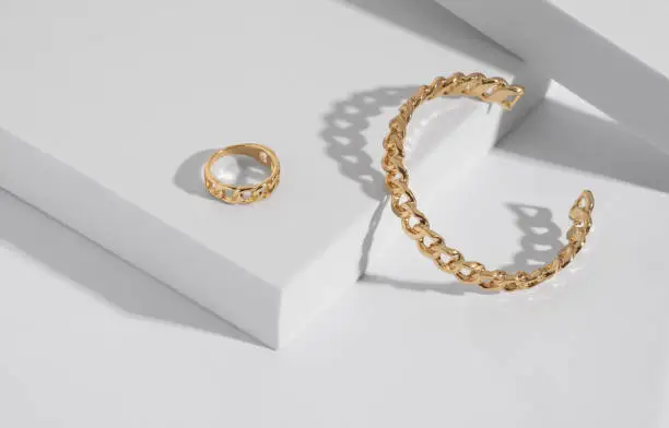 Photo of Chain shape golden modern bracelet and ring on white podium with copy space