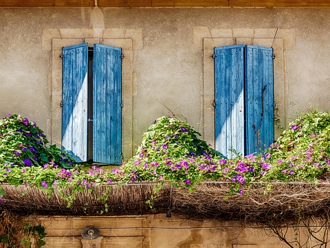 A sunlit balcony is framed by two blue wooden shutters in a scene typical of Provence.