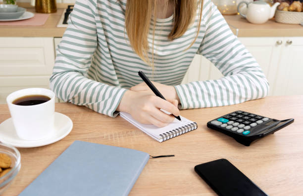 若い女性はノートに書き、電卓で数えます。キッチンの女性がテーブルに座っている。 - teenager problems typing teenagers only ストックフォトと画像