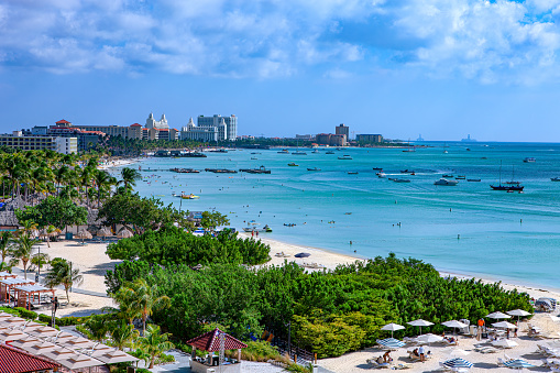 Palm Beach in Aruba on the South Caribbean Sea. Several hotels and boats at anchor are seen in the image. The sea is very calm. In the horizon, is an oil drilling rig. There are a few people in the image but they are too far away to be recognised. Photo shot in the early afternoon sunlight; horizontal format.