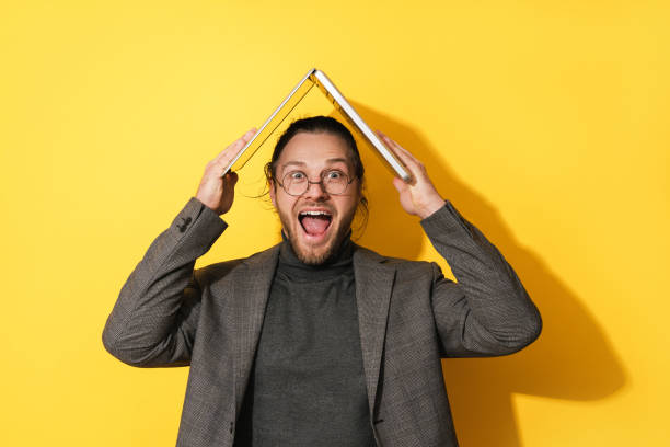 Cheerful bearded man holding laptop computer above his head on yellow background stock photo