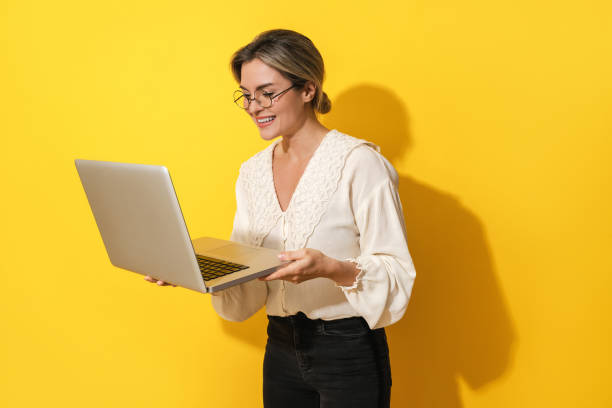 Cheerful woman wearing eyeglasses is using laptop computer on yellow background stock photo