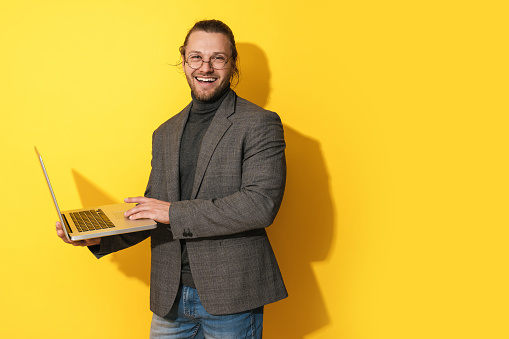 Handsome happy bearded man wearing eyeglasses is using laptop computer on yellow background
