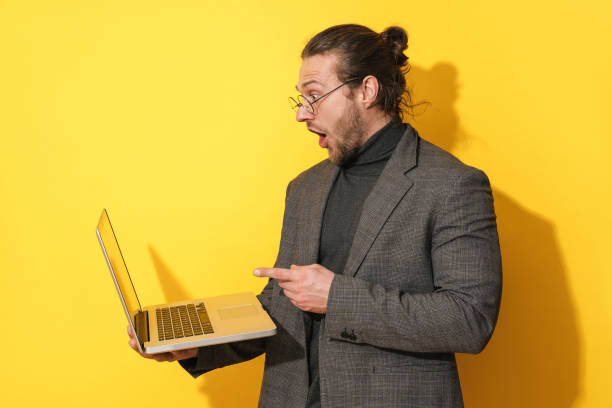 Surprised man wearing eyeglasses pointing at laptop screen on yellow background stock photo