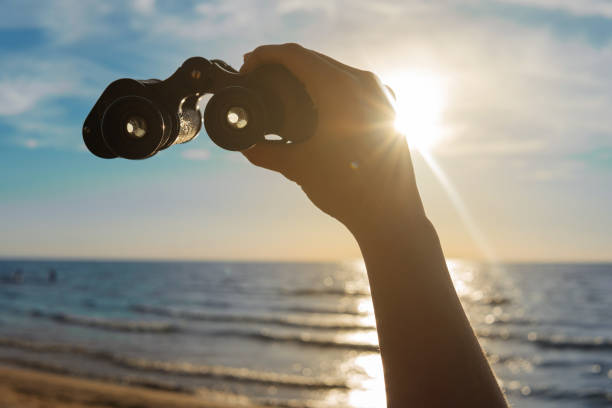 Adventure - Female hand with a vintage binocular against bright sun light on beautiful beach with nice blue sky. stock photo