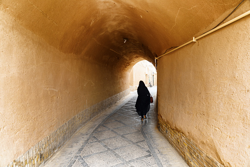 Iranian woman wearing black chador walking along narrow street of the historical city of Yazd. Unique Persian architecture of the ancient town.