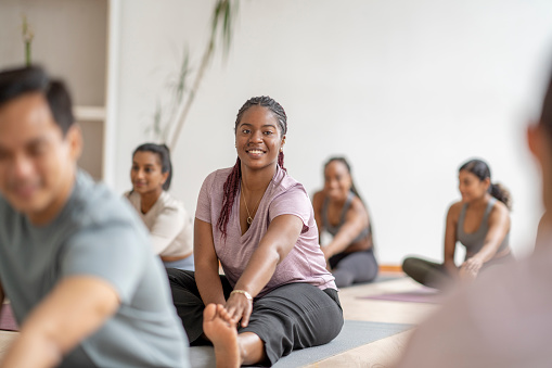 A group of adults sit individually on yoga mats spread around a studio as they warm up their muscles before class.  They are each dresed comfortably in athletic wear and are smiling as they stretch out their legs.