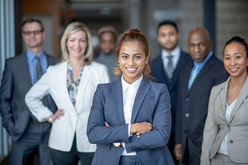 A group of diverse business professionals pose for a group team portrait. The focus is on a middle aged woman with her arms folded across her chest. Each team member is smiling while wearing suits, ties and formal business attire.