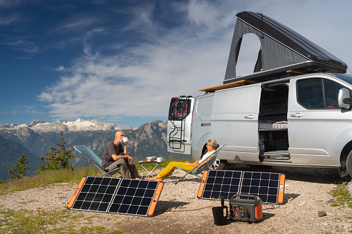 Sporty middle age couple sitting high up in the mountains, dachstein mountain glacier in background, beside their camper van with rooftop tent enjoying breakfast on sunny morning, self sufficient with portable solar panels, part of a series of camping travel