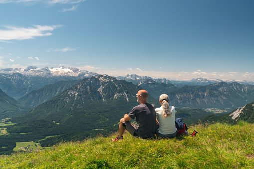 Sporty middle aged couple sitting high up in the mountains in fresh green grass enjoying view at mountain range with dachstein glacier in austria on sunny day, rear view and place for text, part of a series of camper van travel