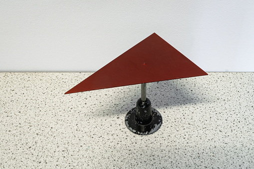 Physics experiment where a red triangular metal plate is balancing on a needle. The center of gravity of the triangle is placed exactly at the tip of the needle.