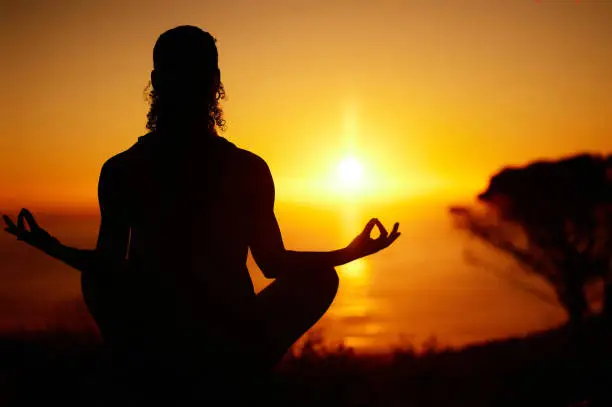 Photo of Rearview silhouette yoga woman meditating with legs crossed for outdoor practice in remote nature. Mindful person sitting alone and balancing for mental health at sunset. Serene and zen in lotus pose