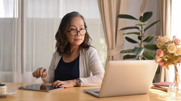 istock Mature woman watching online webinar on laptop and using digital tablet during remote working from home. 1403603126