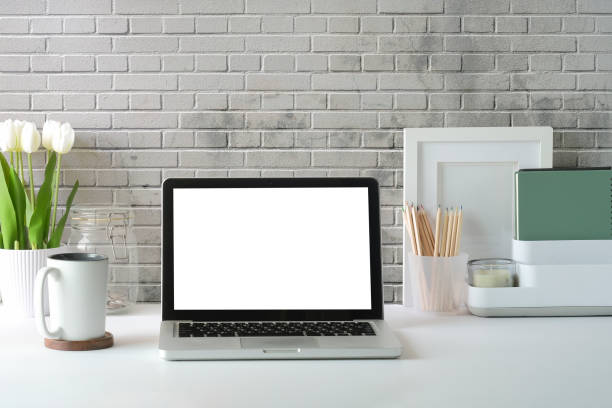istock Computer laptop with blank screen, flower pot, coffee cup and picture frame on white table. 1403609839