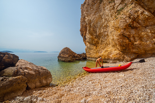 Sporty mature woman preparing her kayak on a beautiful stone beach between rocks and cliffs to start paddleing in clear waters of croatian sea